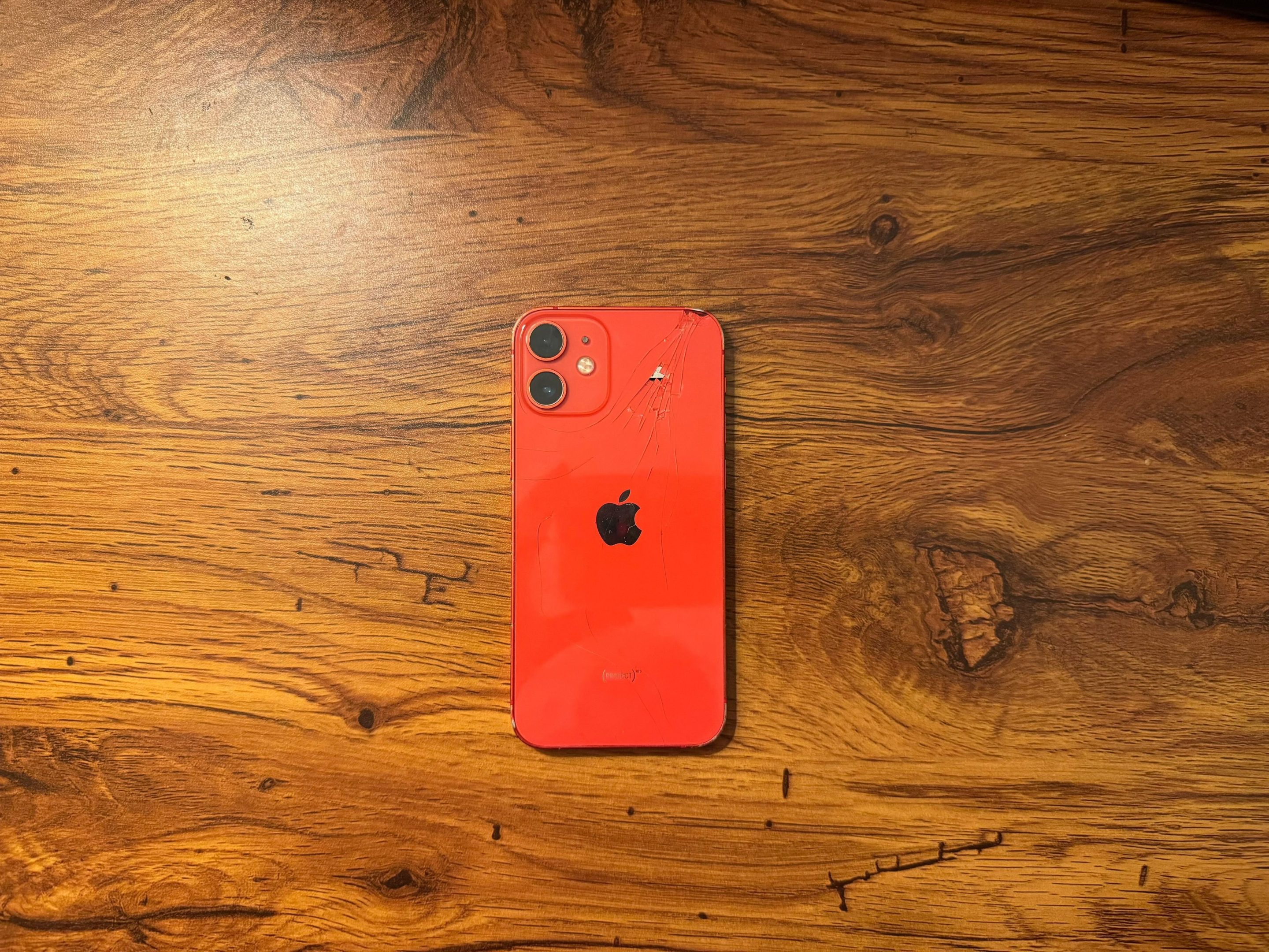 A red iPhone 12 mini rests on a wood table. The back of the phone is facing up; it's glass surface is cracked.