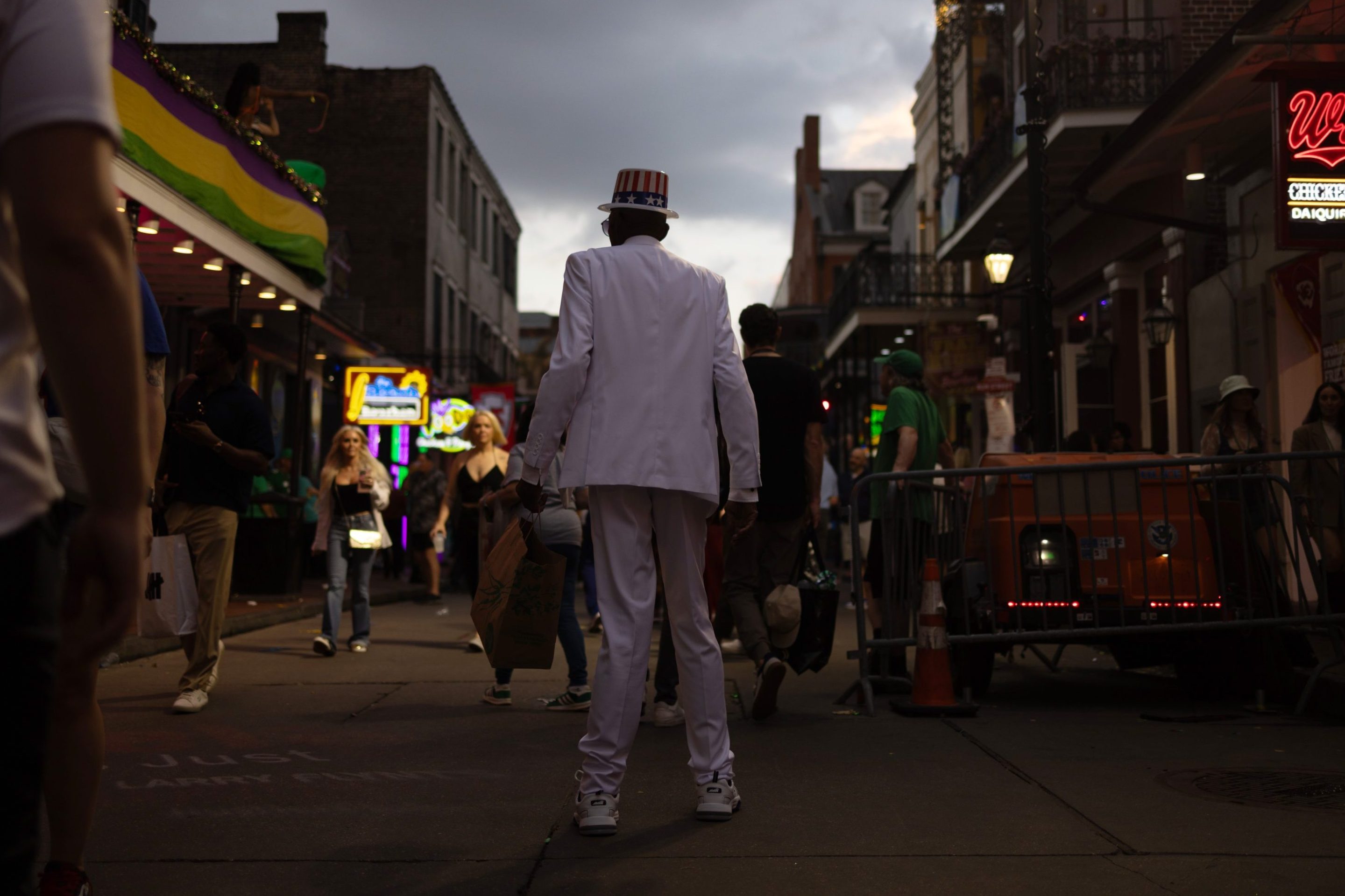 A photo of a man on stilted legs and dressed in white suit walking down a dark and mostly empty Bourbon Street in New Orleans.