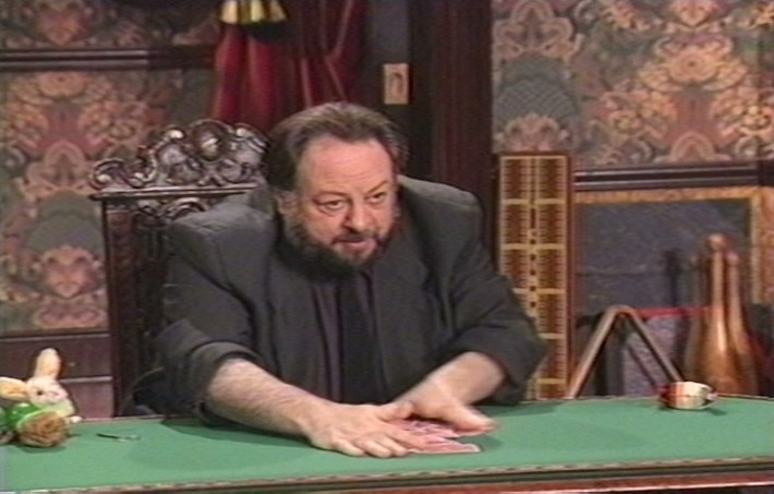 Ricky Jay, with his hair balding halfway up his head but black and tall, and a large black beard, in a black suit with a black tie and a dark purple shirt, looking at the camera at a poker table shuffling cards on it.