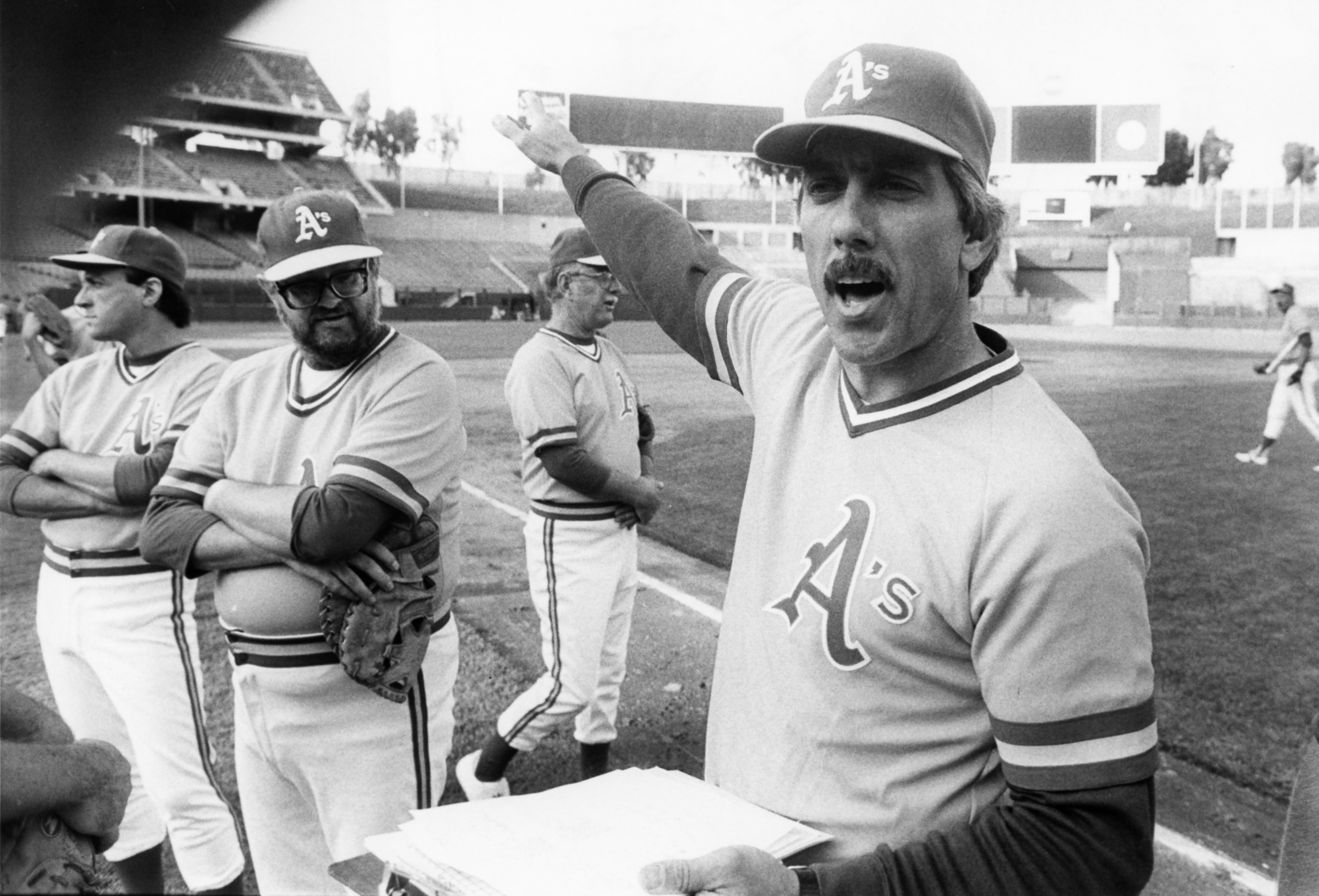 A black-and-white image of Ray Fosse, in an old-timey Oakland A's uniform, bossing around a bunch of lumpy '70s guys at an A's fantasy camp in