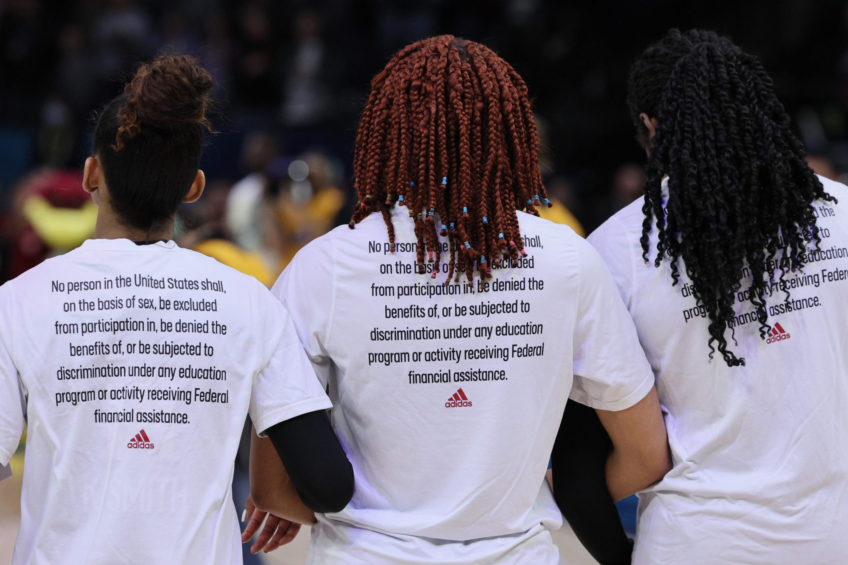 A photo of three women's basketball players. They all stand with their backs to the camera. On the back of their shirts are the 37 words that make up Title IX, the law that gave women equal access to school sports