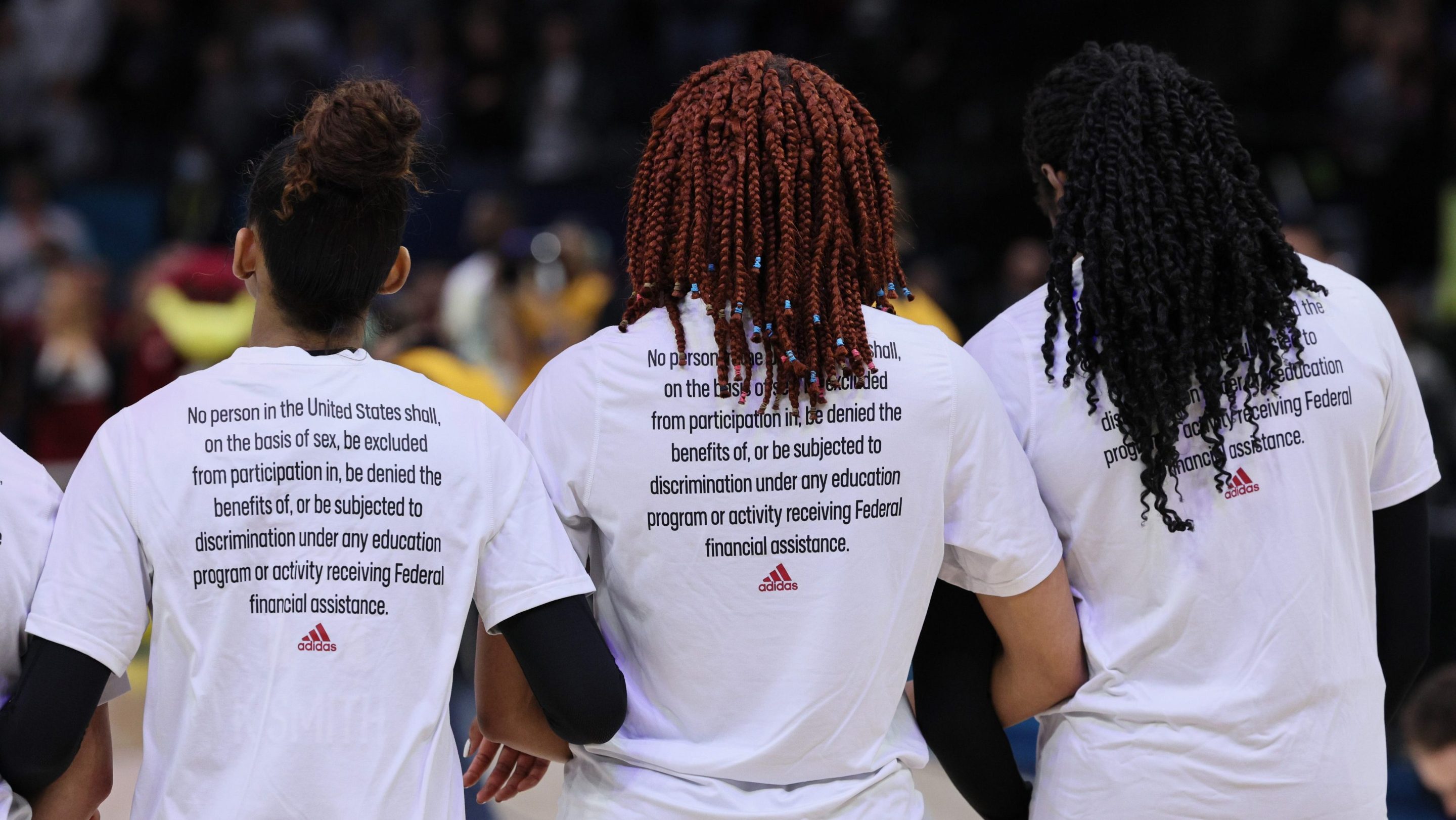 A photo of three women's basketball players. They all stand with their backs to the camera. On the back of their shirts are the 37 words that make up Title IX, the law that gave women equal access to school sports