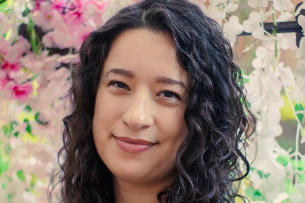a photo of Sabrina Valenti, formerly a budget analyst for NOAA. Sabrina has brown curly hair and is standing against a floral background.