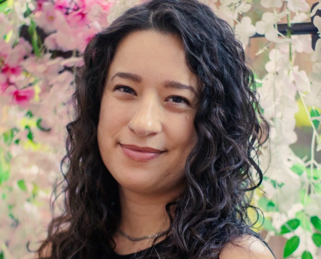 a photo of Sabrina Valenti, formerly a budget analyst for NOAA. Sabrina has brown curly hair and is standing against a floral background.