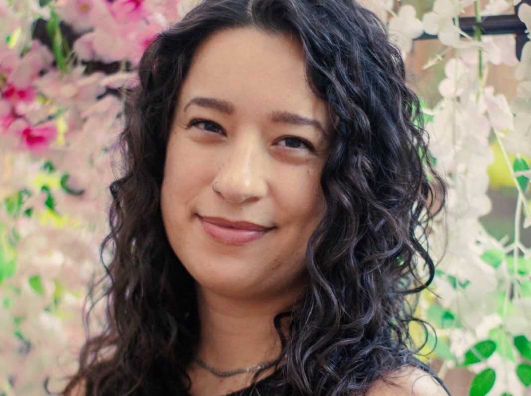 a photo of Sabrina Valenti, formerly a budget analyst for NOAA. Sabrina has brown curly hair and is standing against a floral background.