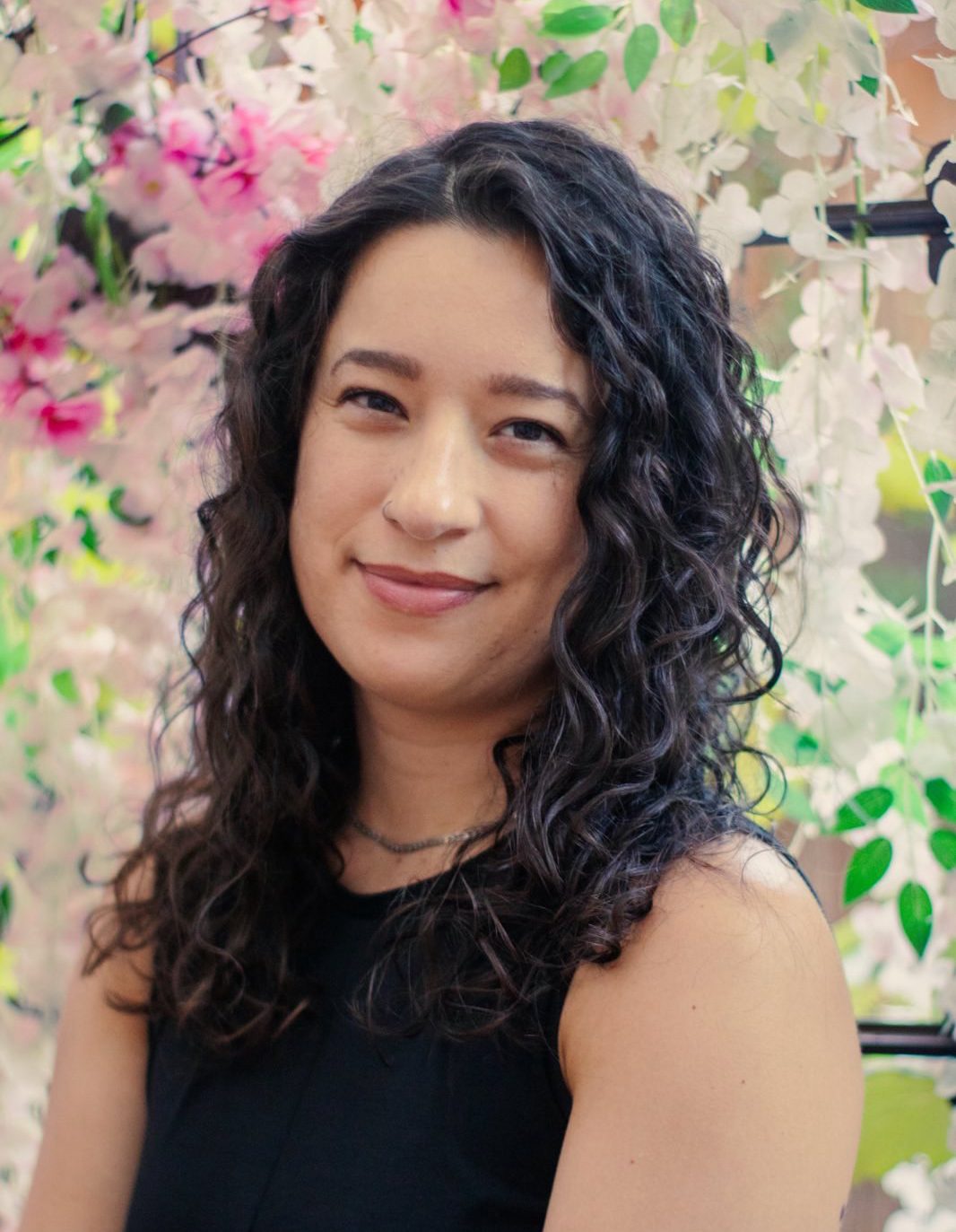 a photo of Sabrina Valenti, formerly a budget analyst for NOAA. Sabrina has brown curly hair and is standing against a floral background.