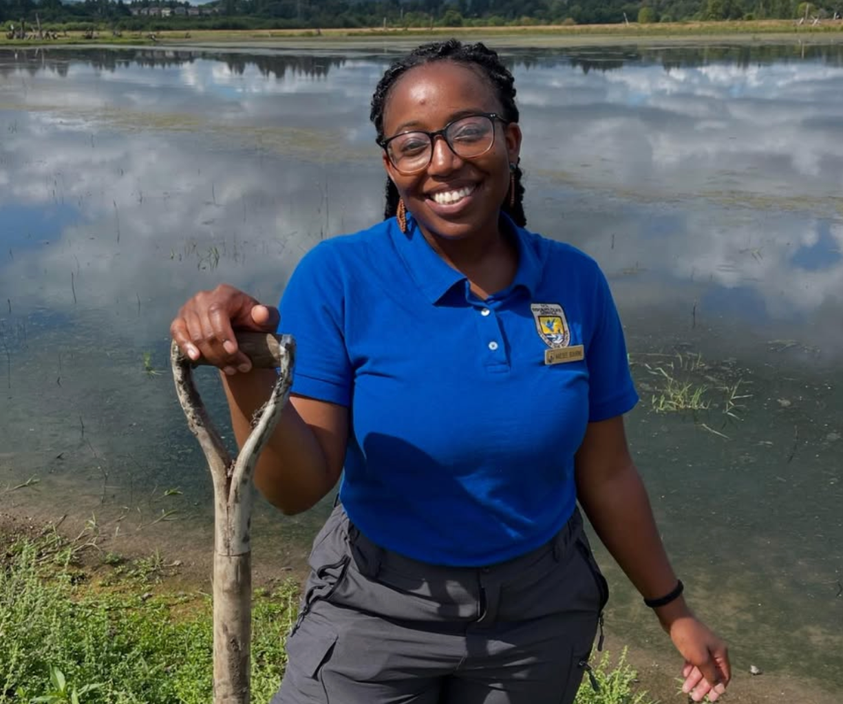 a photo of Kaesee Bourne, a former fish and wildlife biologist at the USFWS, holding a shovel in front of a wetland