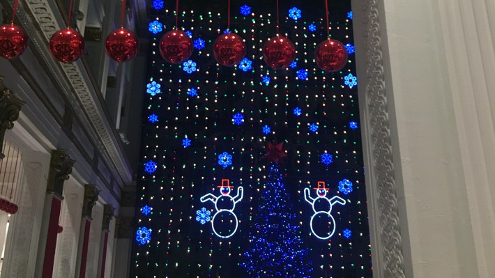 A wall of lights goes up the inside of a building. A Christmas tree lit in blue is in the center, surrounded by two light-up snowmen. Snowflakes, made by lights, go up the wall of lights. The shot is from far away, with a wall/column on the right side of the photo.