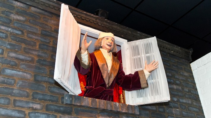 A character, animatronic-esque, on the second floor of a building leaning out a window where both sides have been pushed open. The old man, in a robe and wearing a stocking cap, is Ebenezer Scrooge on Christmas Day.