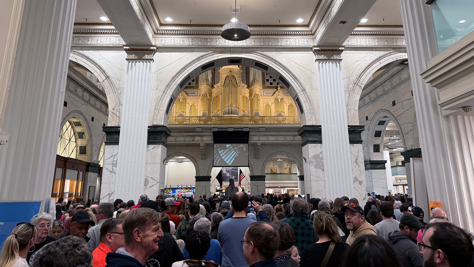 Photo of a white marble room with three archways in the middle. In the middle archway is a giant gold pipe organ, with two more archways on the other side of the room. A huge crowd fills the bottom of the photo, listening to the organ.