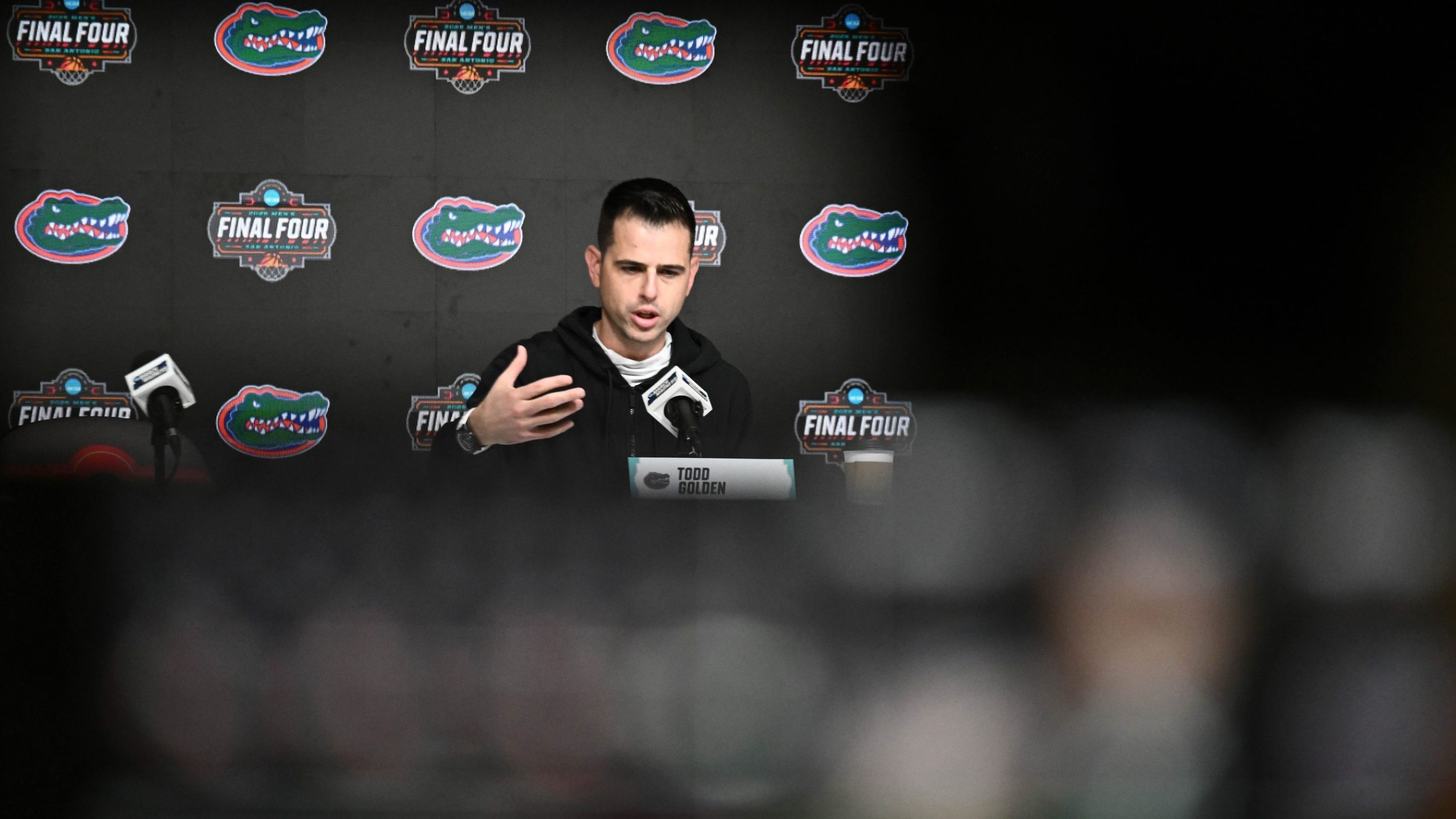 A photo of Florida Gators basketball coach Todd Golden speaking to reporters at a press conference during the Final Four. Golden is sitting at a table with a nameplate.