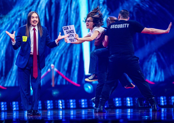 Tommy Cash, in a suit with a long red tie, performs at Eurovision 2025. Two fake security members are holding back a fake fan with a "CAN I DANCE WITH YOU" sign.