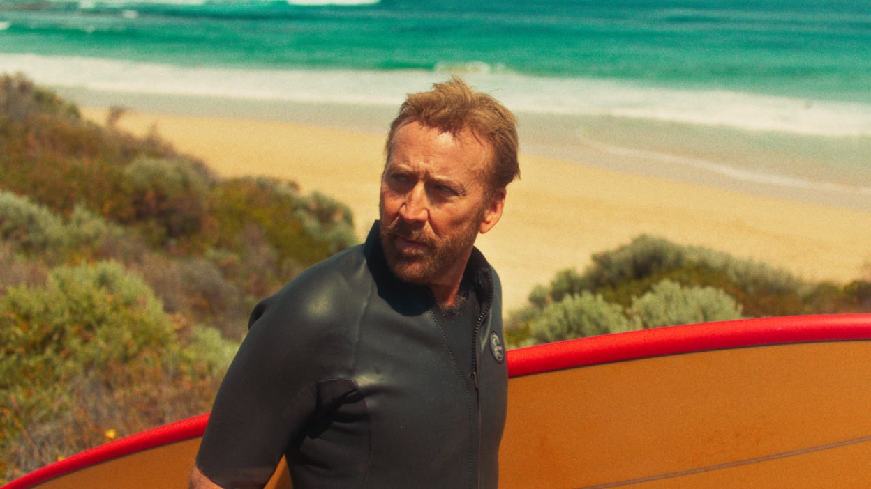 Nicolas Cage on a beach holding a surfboard.
