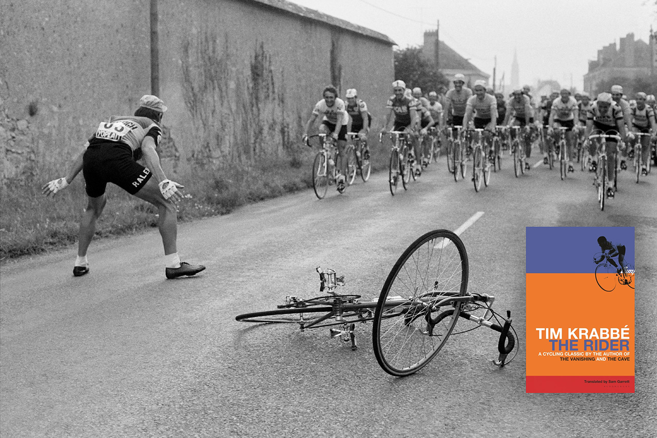 Cyclists in a race are in the background. There is a house or wall on the left side of the road. There is a man in front, looking at the peloton, squatting halfway with his arms spread just past his knees. Next to him in the center of the road is a bicycle laid across the ground.