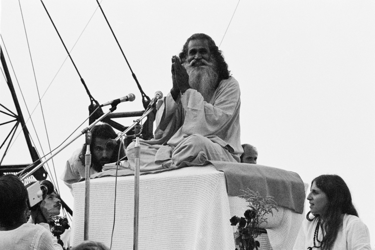 Swami Satchidananda giving the opening speech at the Woodstock Music Festival, Bethel, New York, 15th August 1969. He is sitting atop a raised seat and speaking into several mics. His legs are crossed and his hands are together in a prayer-like shape.