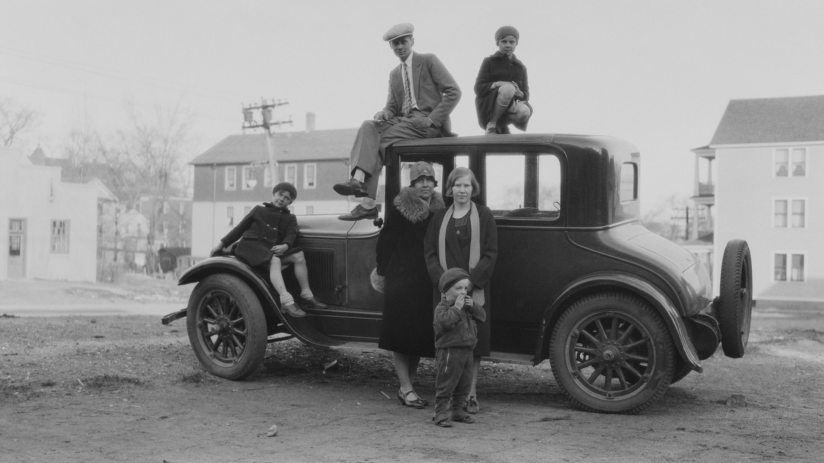 A family group shot of three children, two women and a cigarette smoking man gathered around and standing on top of a car roof parked on an wide street, several houses in the background, America, circa 1925.