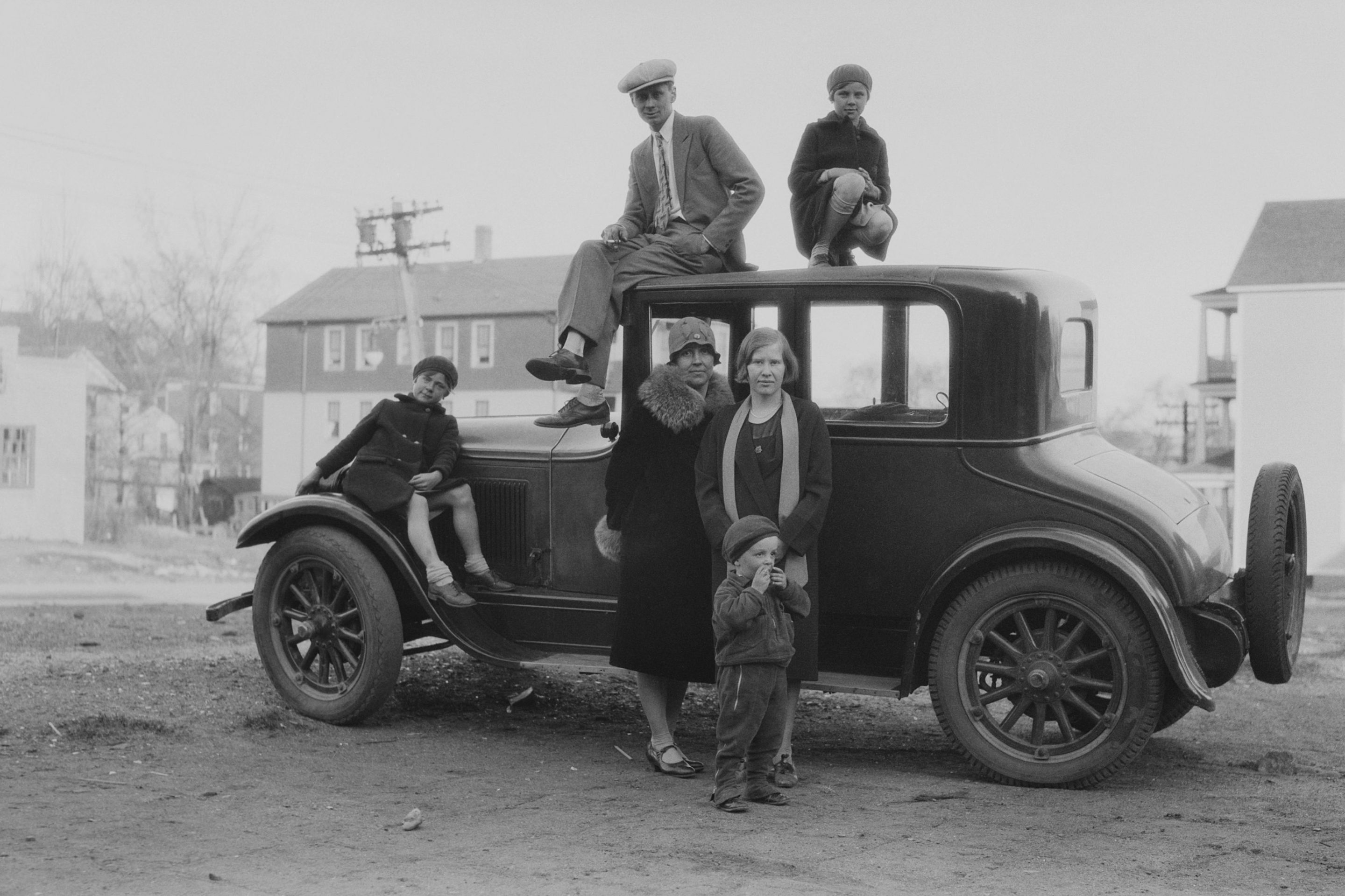 A family group shot of three children, two women and a cigarette smoking man gathered around and standing on top of a car roof parked on an wide street, several houses in the background, America, circa 1925.
