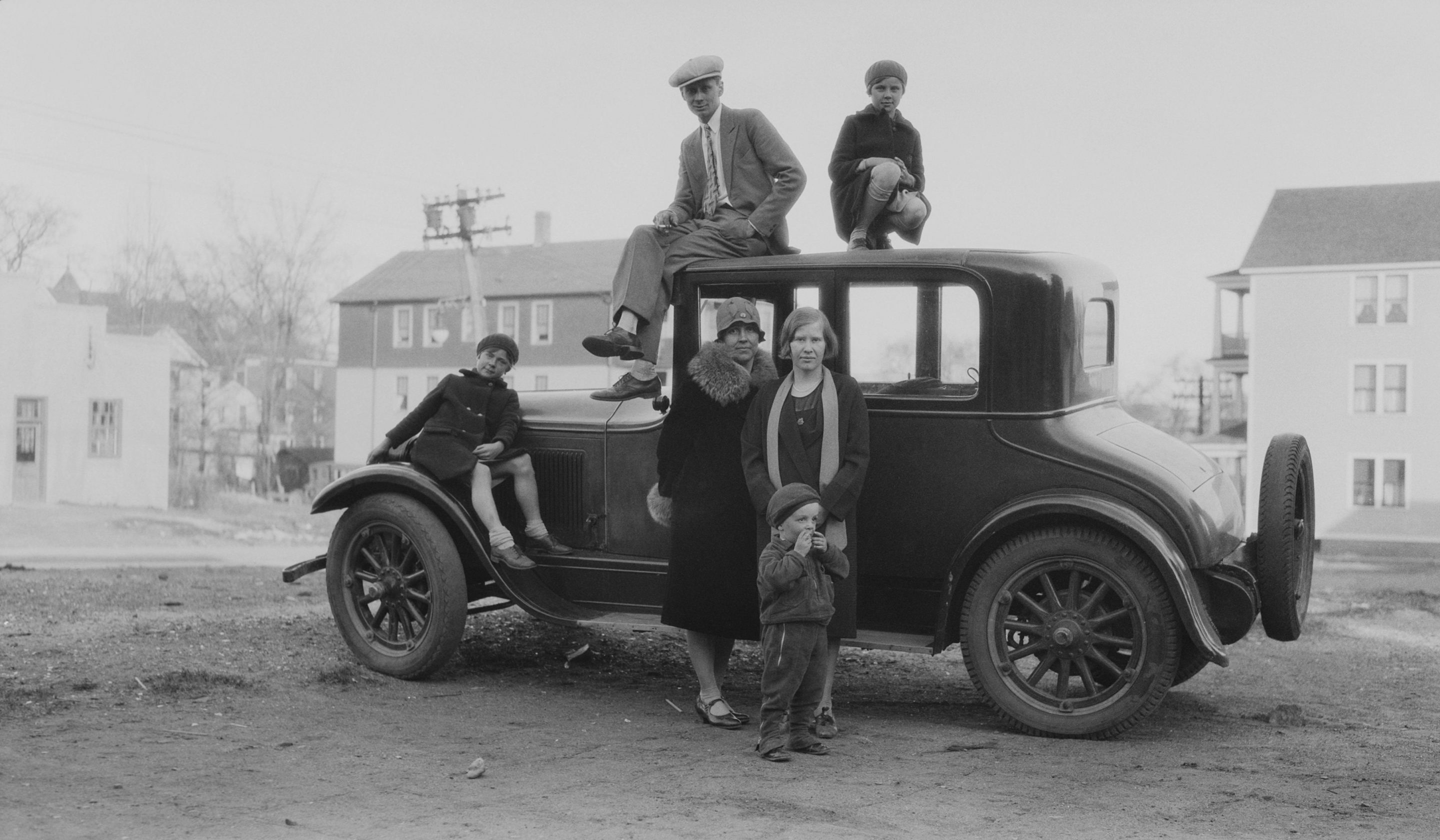 A family group shot of three children, two women and a cigarette smoking man gathered around and standing on top of a car roof parked on an wide street, several houses in the background, America, circa 1925.