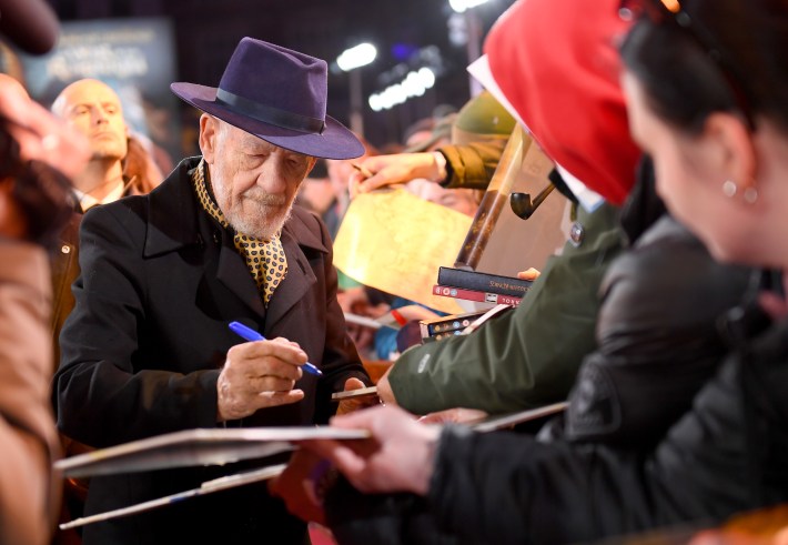 Ian McKellen attends "The Lord Of The Rings: The War of The Rohirrim" World Premiere at the Odeon Luxe Leicester Square on December 03, 2024 in London, England. He is signing autographs for fans, wearing a coat, a tie, and a purple hat.