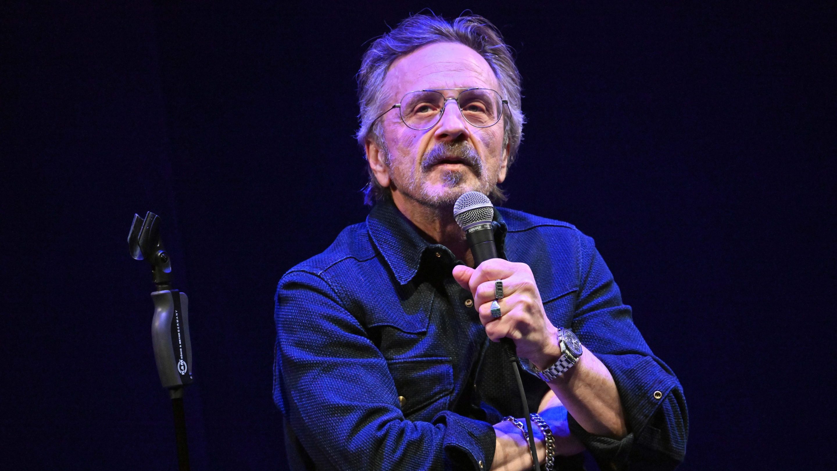Marc Maron performing stand up comedy. He is sitting on a stool and holding a microphone