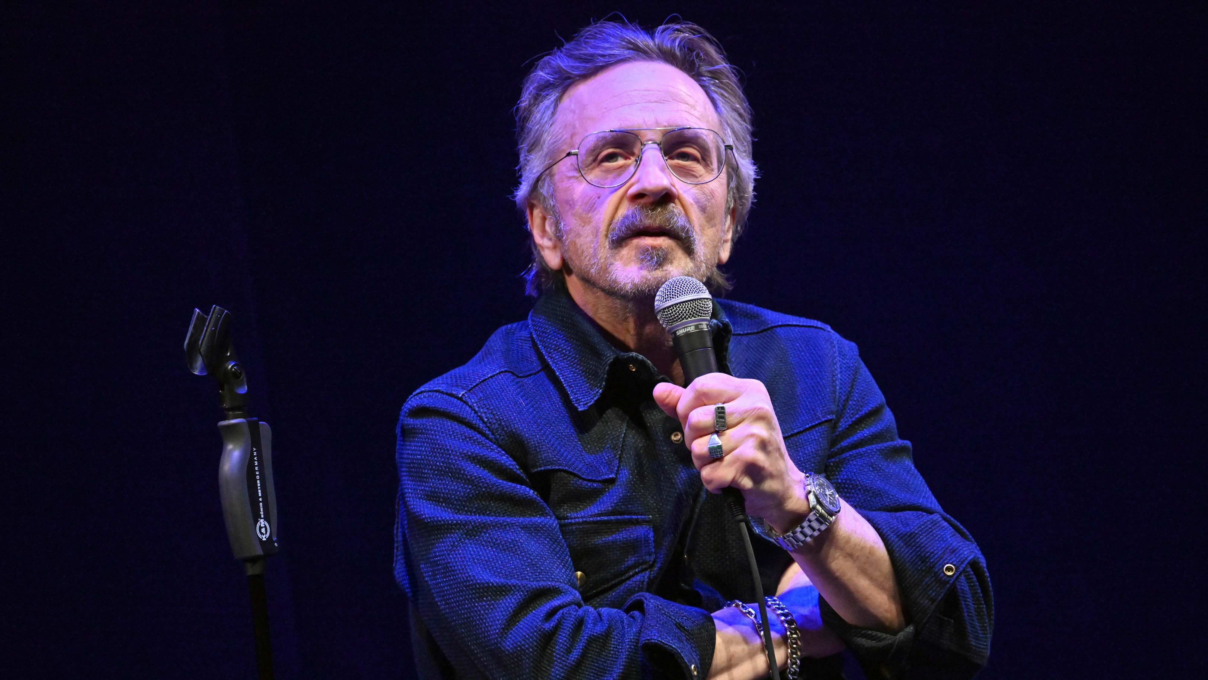Marc Maron performing stand up comedy. He is sitting on a stool and holding a microphone
