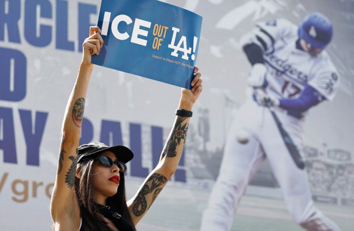 A protestor holds up a sign that says "ICE out of LA!" in front of a billboard advertising the Dodgers.