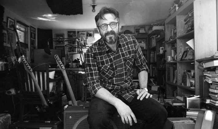 A photo of Marc Maron sitting in his garage. It shows behind him guitars, boxes, and a lot of books. He's wearing his glasses, pants, and a flannel. The photo is in black and white.