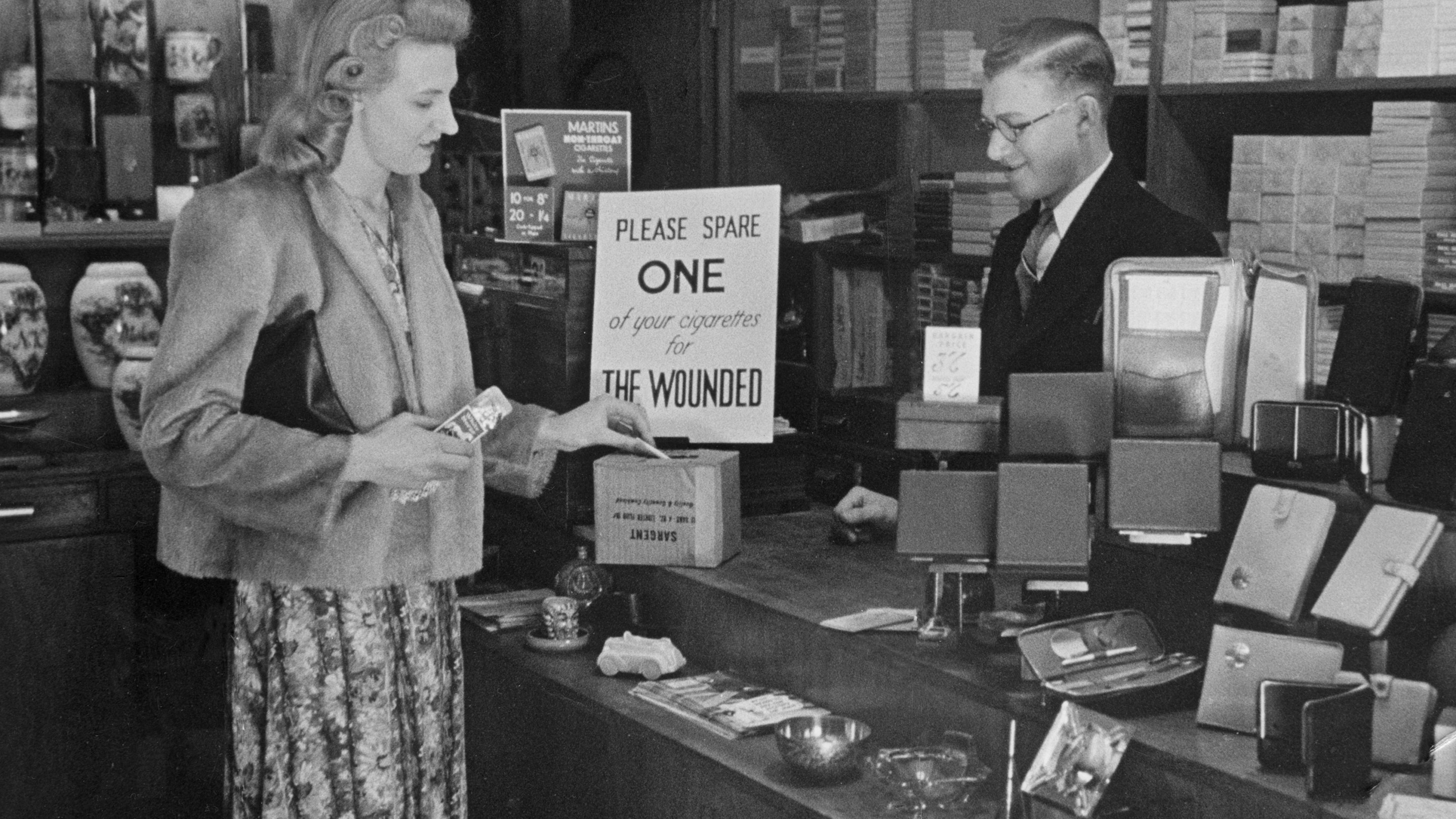A woman buying a packet of cigarettes in a tobacconist shopin London, leaves a spare one in a cigarette collection box, which will be sent on to a hospital for wounded soldiers during the Second World War June 1940
