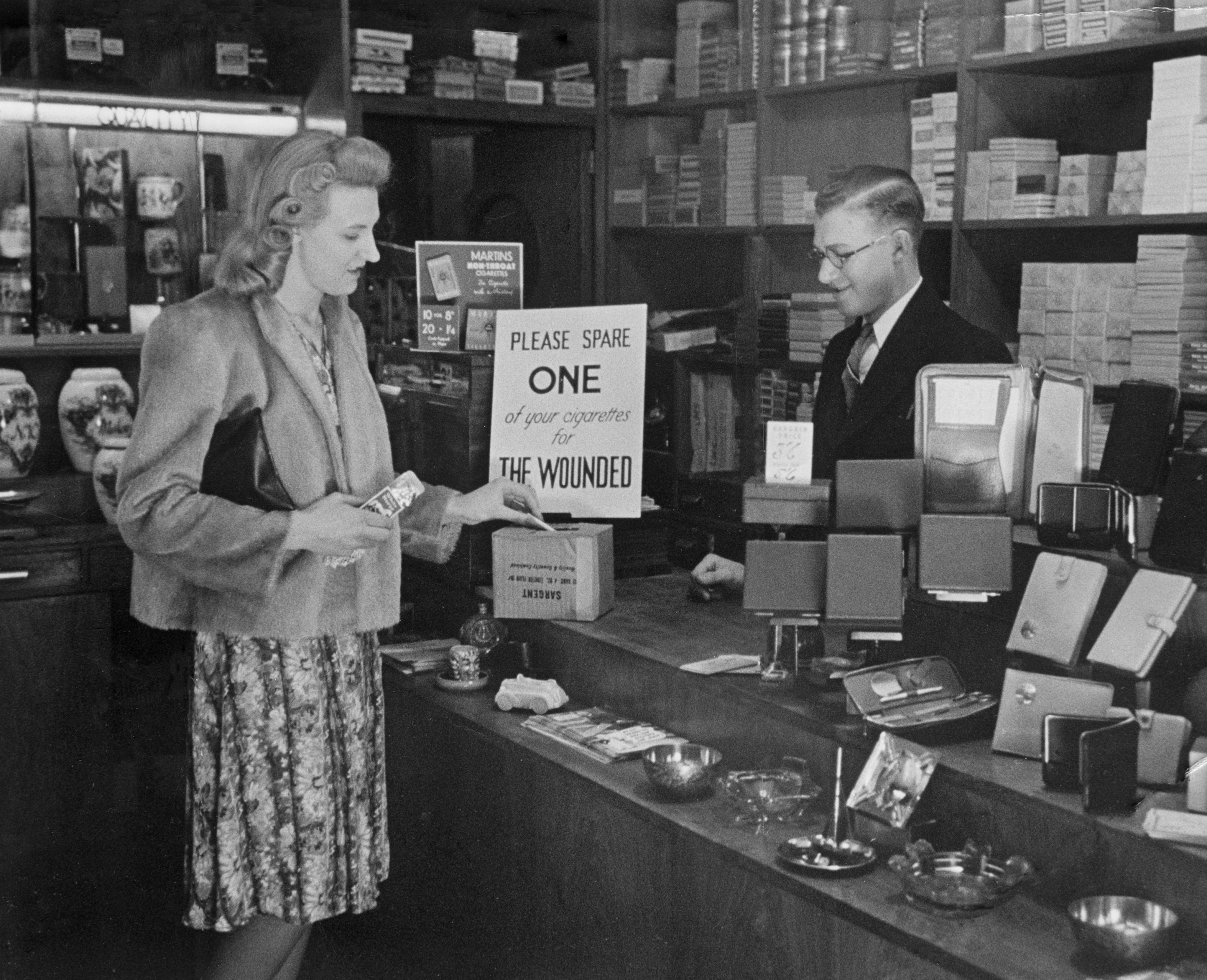 A woman buying a packet of cigarettes in a tobacconist shopin London, leaves a spare one in a cigarette collection box, which will be sent on to a hospital for wounded soldiers during the Second World War June 1940