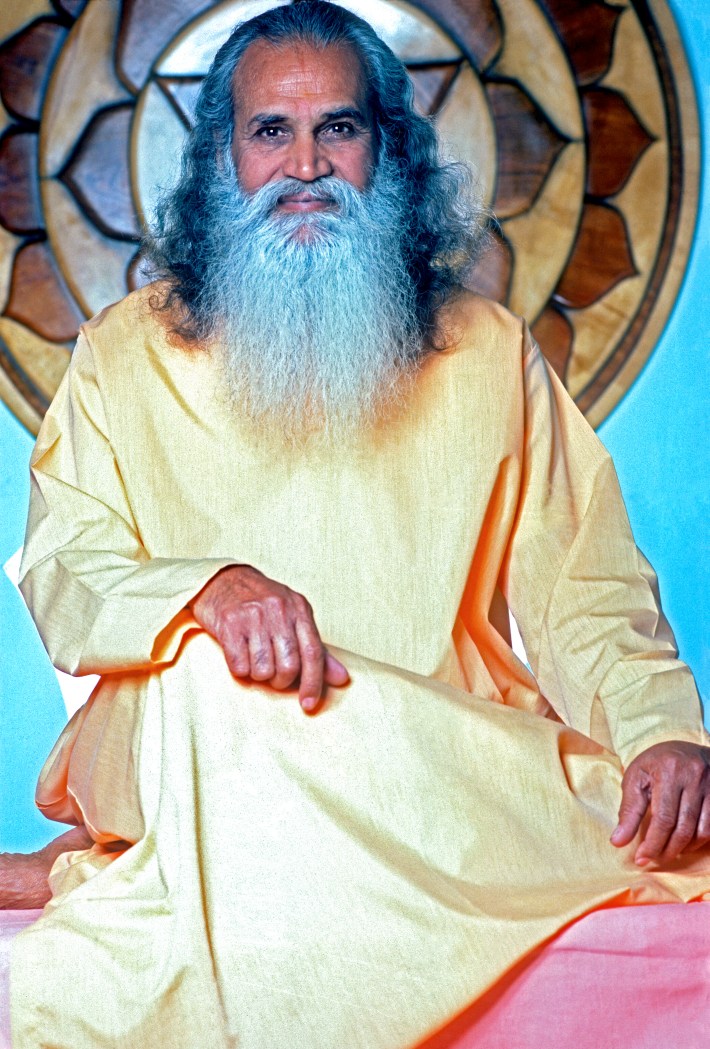 Swami Satchidananda poses for a portrait circa December, 1976 at the IYI Institue in San Francisco, California. He is sitting cross-legged, wearing a yellow rob, and slightly smiling.