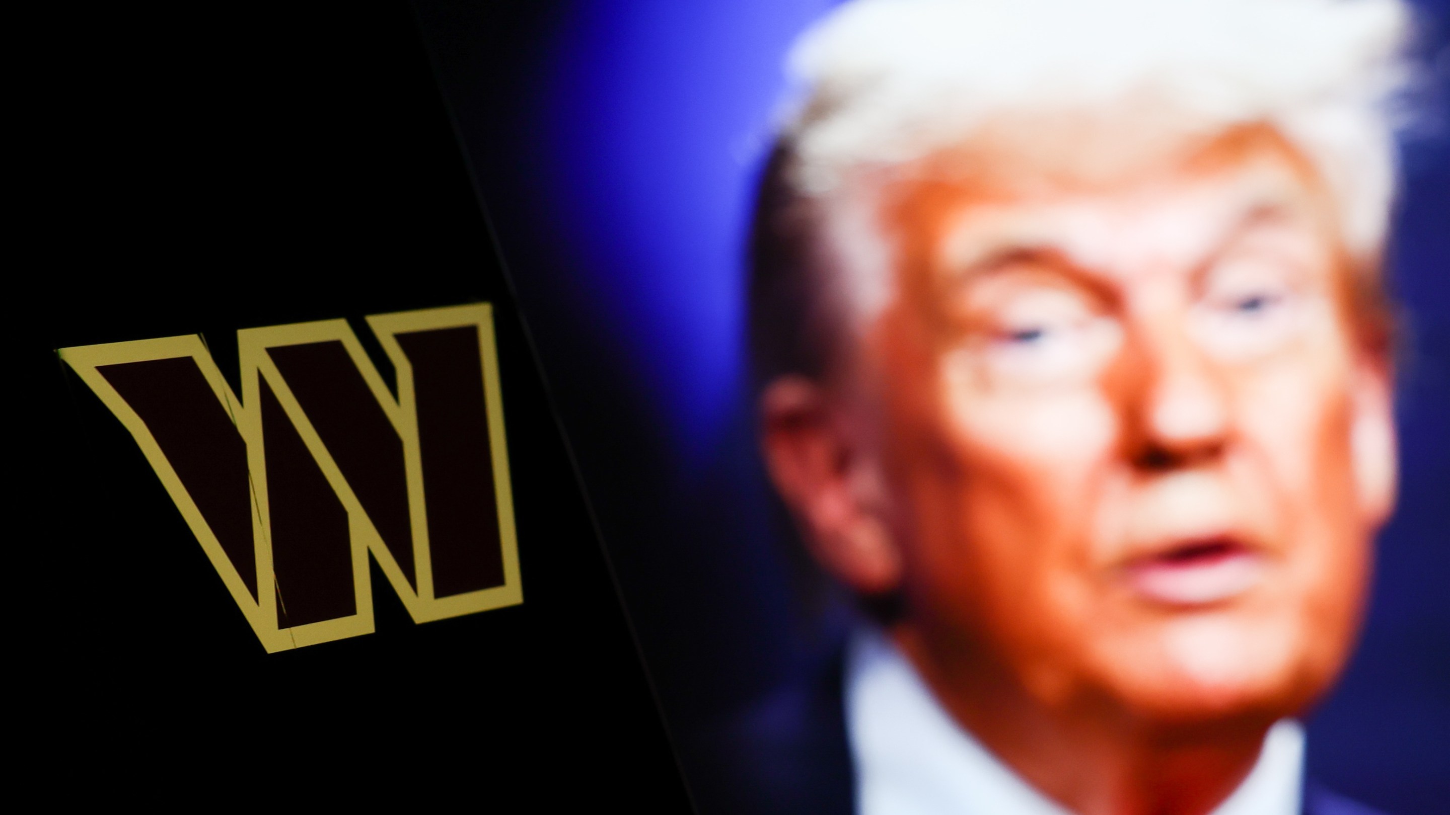Washington Commanders logo displayed on a phone screen and photo of Donald Trump taken on June 25 in The Hague displayed on a laptop screen in the background. It's a photo illustration.