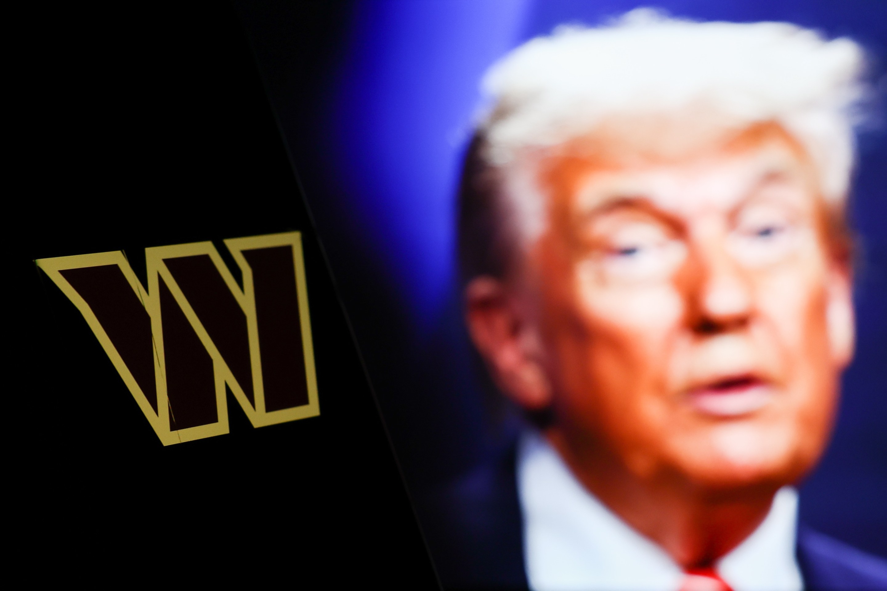 Washington Commanders logo displayed on a phone screen and photo of Donald Trump taken on June 25 in The Hague displayed on a laptop screen in the background. It's a photo illustration.
