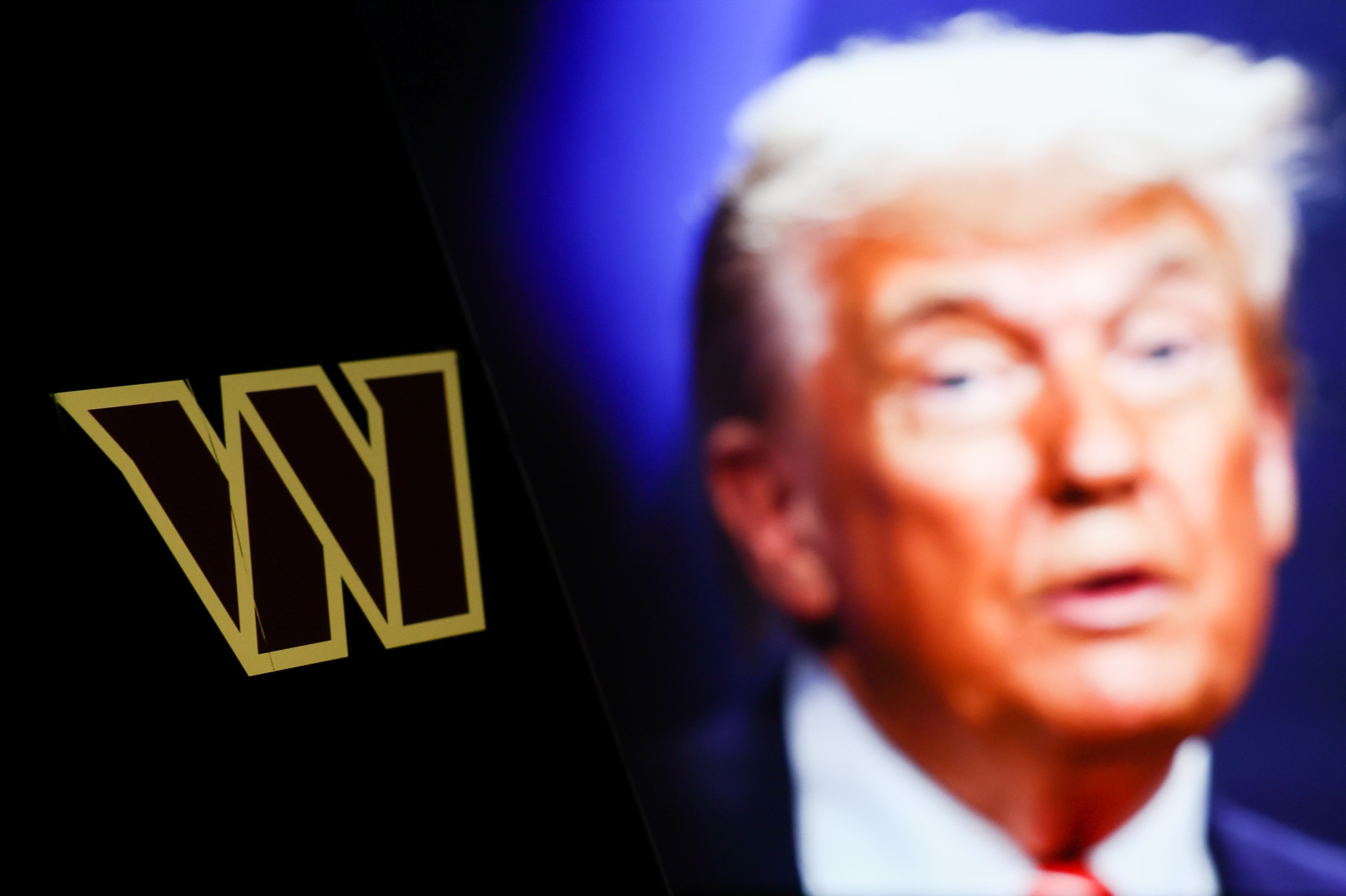 Washington Commanders logo displayed on a phone screen and photo of Donald Trump taken on June 25 in The Hague displayed on a laptop screen in the background. It's a photo illustration.