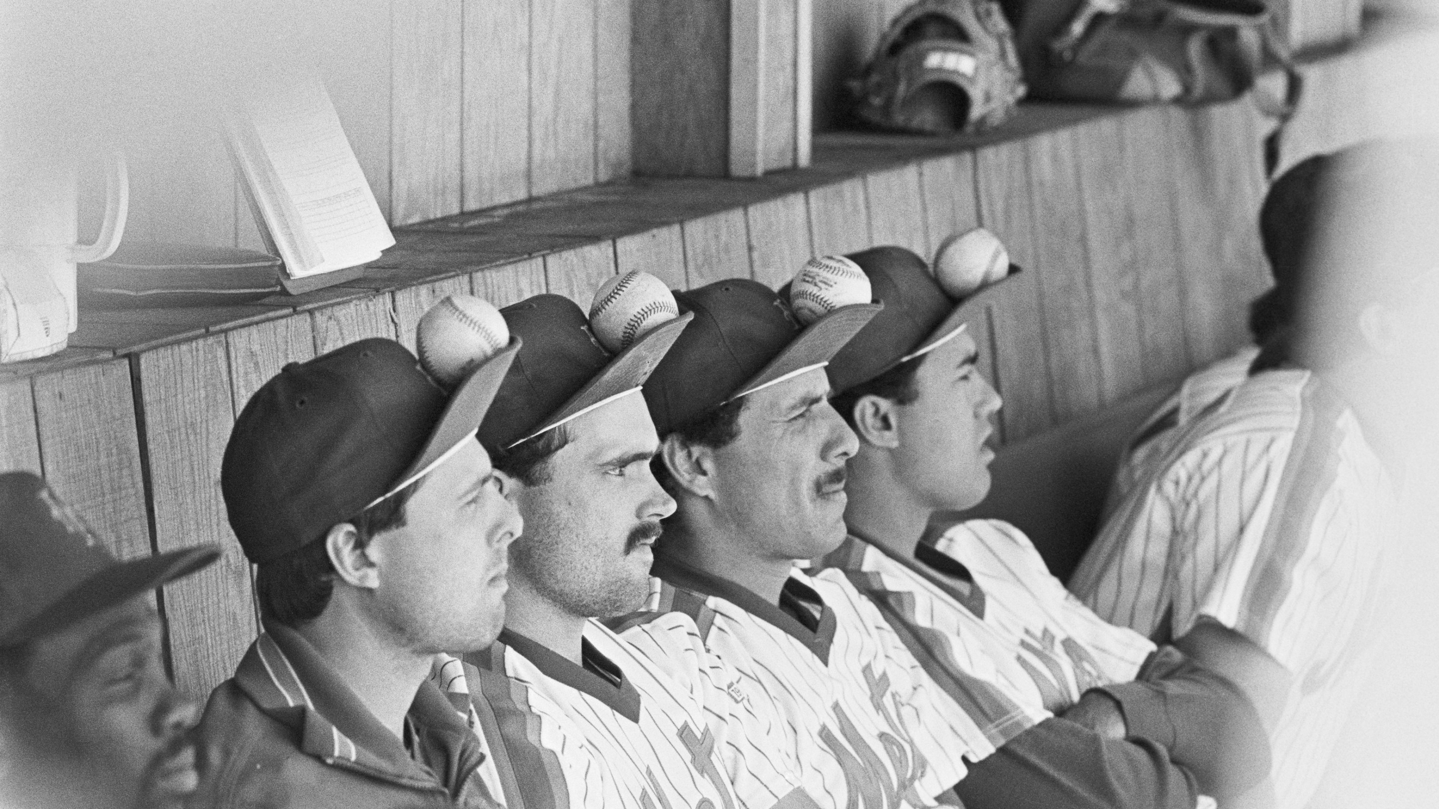 Sitting in the dugout and watching the game, (L to R), are Tim Teufel, Howard Johnson, Bob Ojeda and Ron Darling, as they balance baseballs on their caps to try and change the Mets luck. The image is in black and white; and from 1986.
