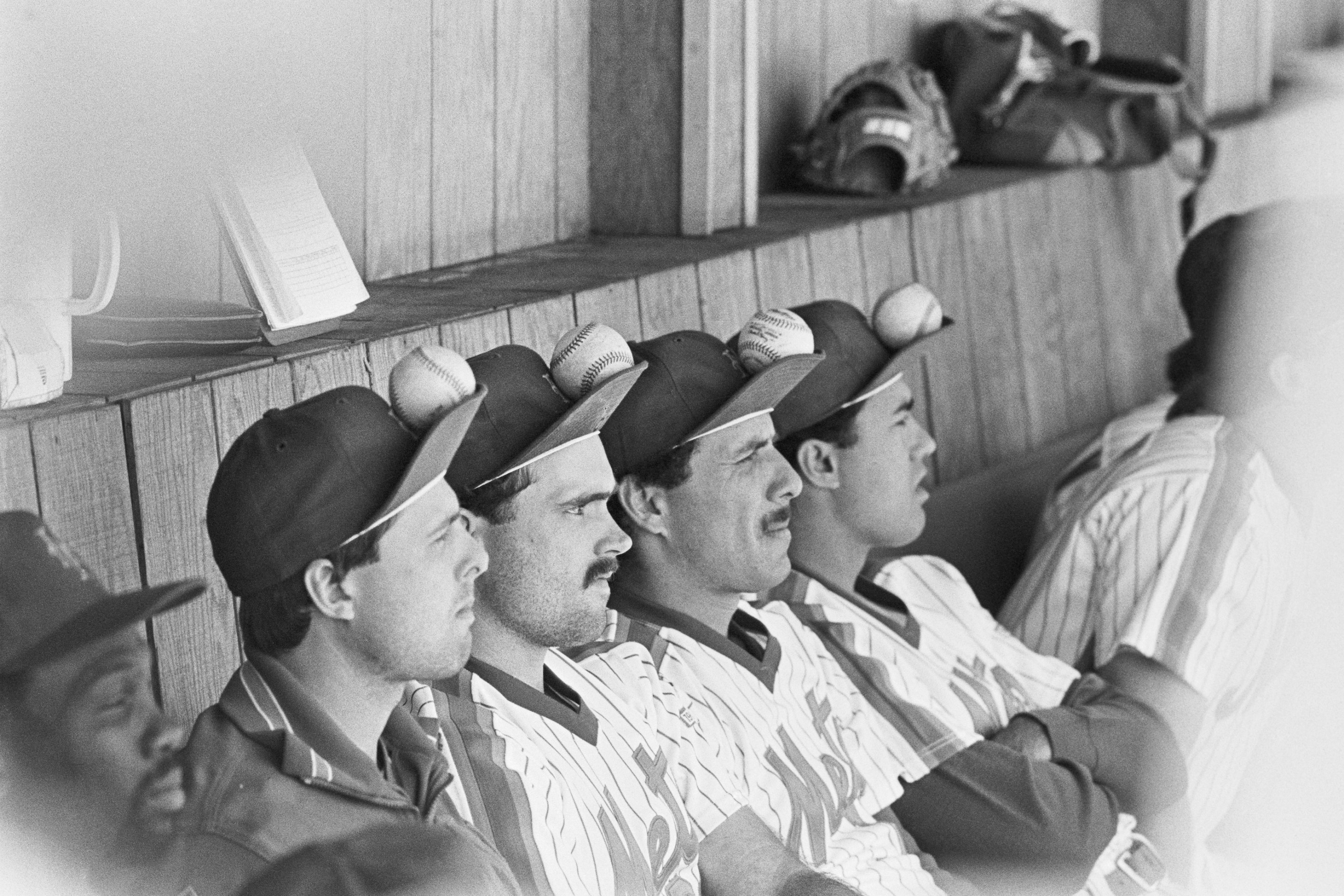 Sitting in the dugout and watching the game, (L to R), are Tim Teufel, Howard Johnson, Bob Ojeda and Ron Darling, as they balance baseballs on their caps to try and change the Mets luck. The image is in black and white; and from 1986.