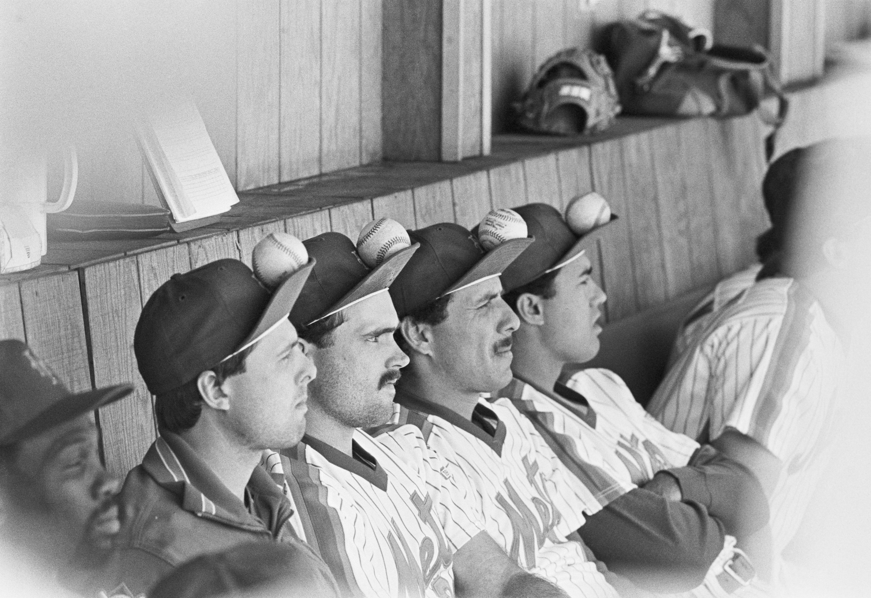 Sitting in the dugout and watching the game, (L to R), are Tim Teufel, Howard Johnson, Bob Ojeda and Ron Darling, as they balance baseballs on their caps to try and change the Mets luck. The image is in black and white; and from 1986.