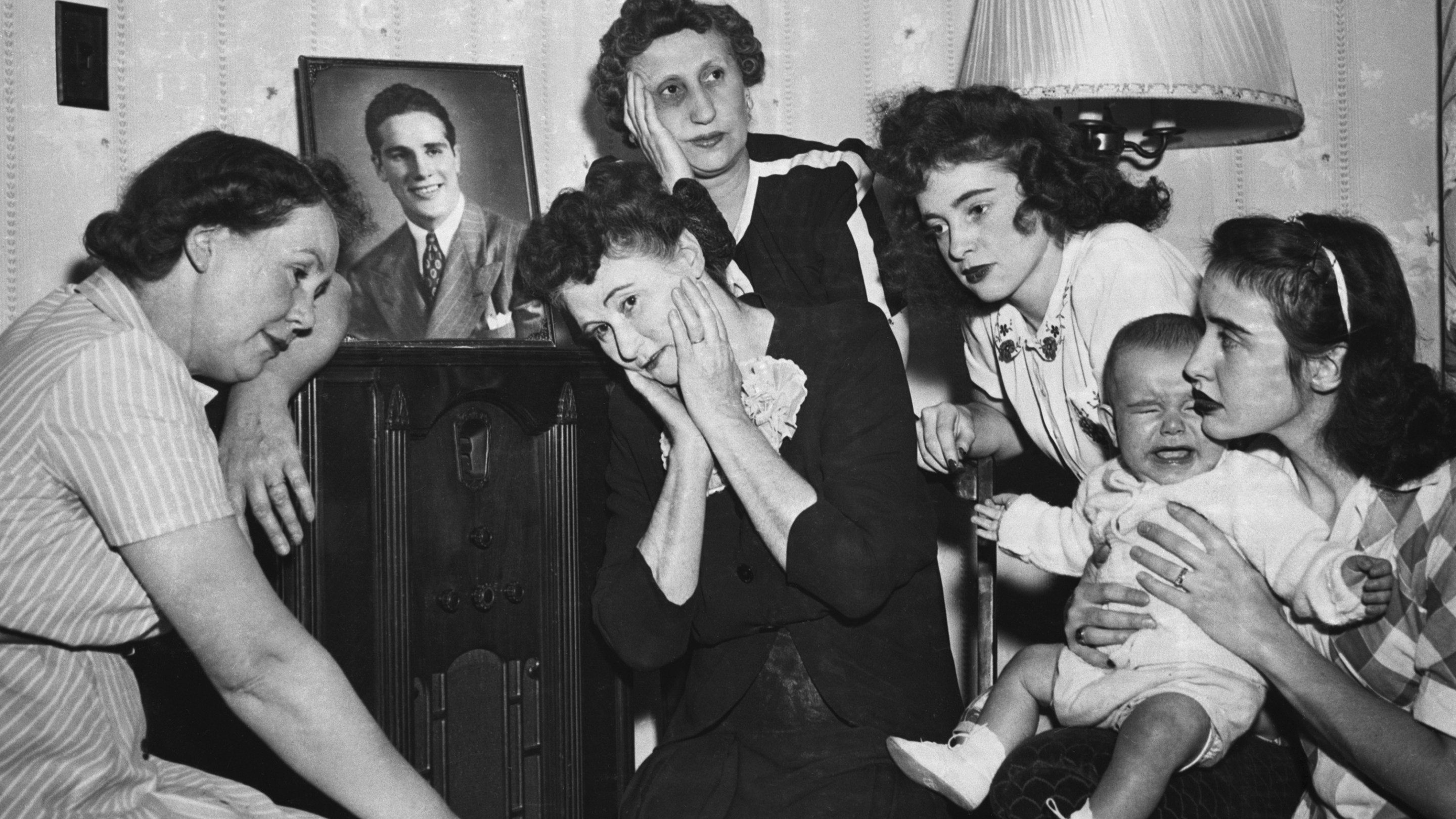 A group of people gathers around an old timey radio to listen to a boxing match.