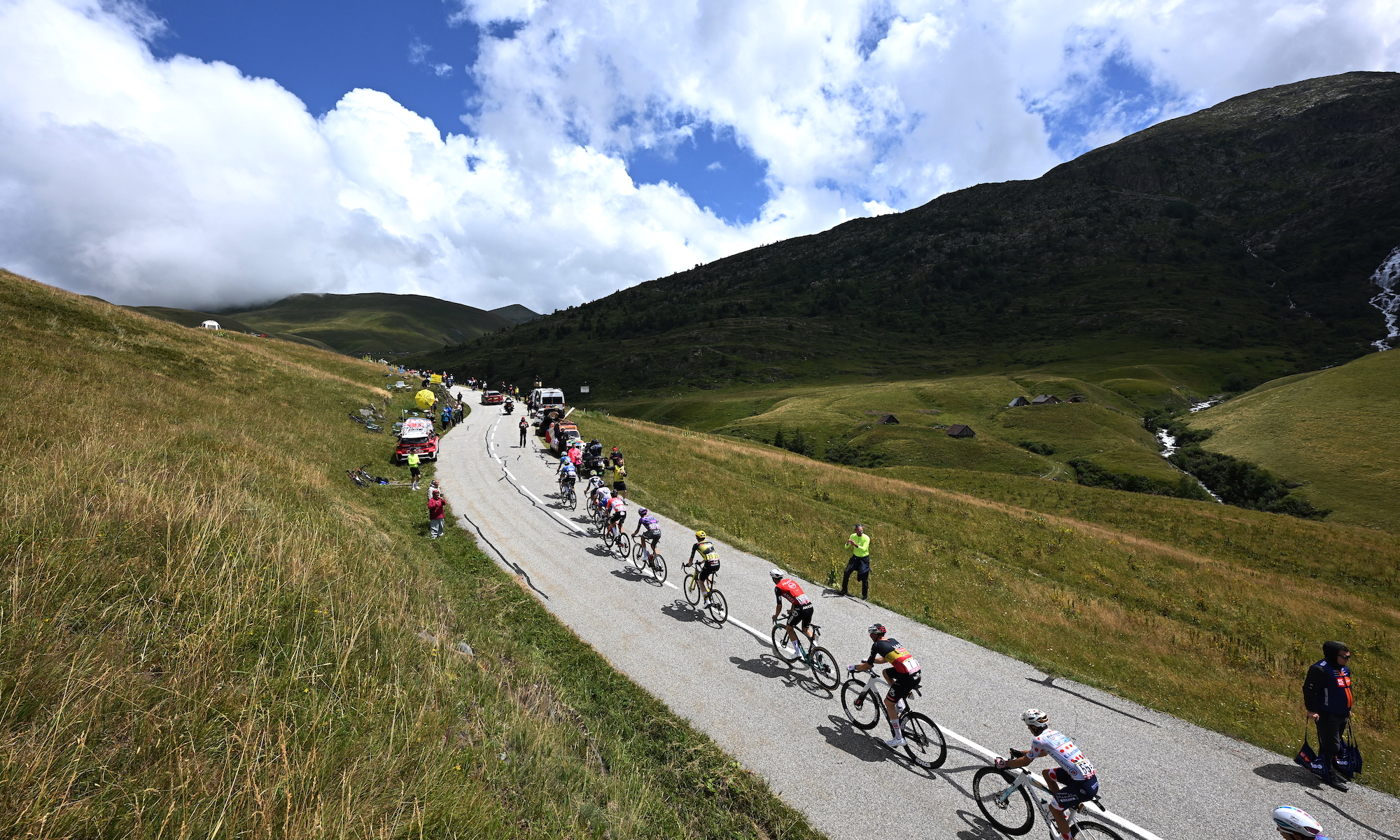 Tour de France riders against a lovely mountain backdrop dappled with the shadows of fluffy clouds.