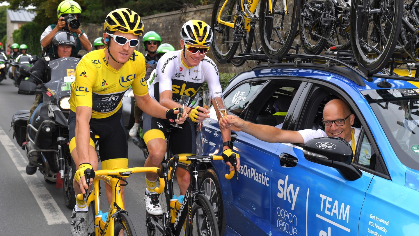 Team Sky cyclist clinks champagne glasses with team car driver while riding during 2018 Tour de France.