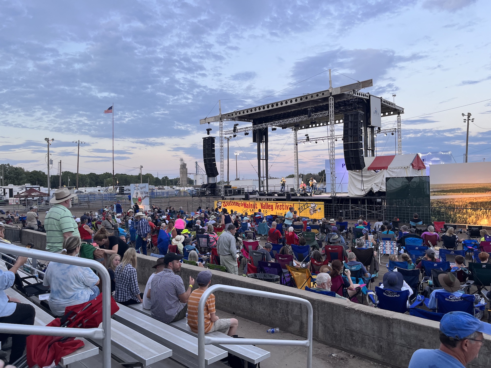 People sit in bleachers and lawn chairs in front of a stage at sunset.
