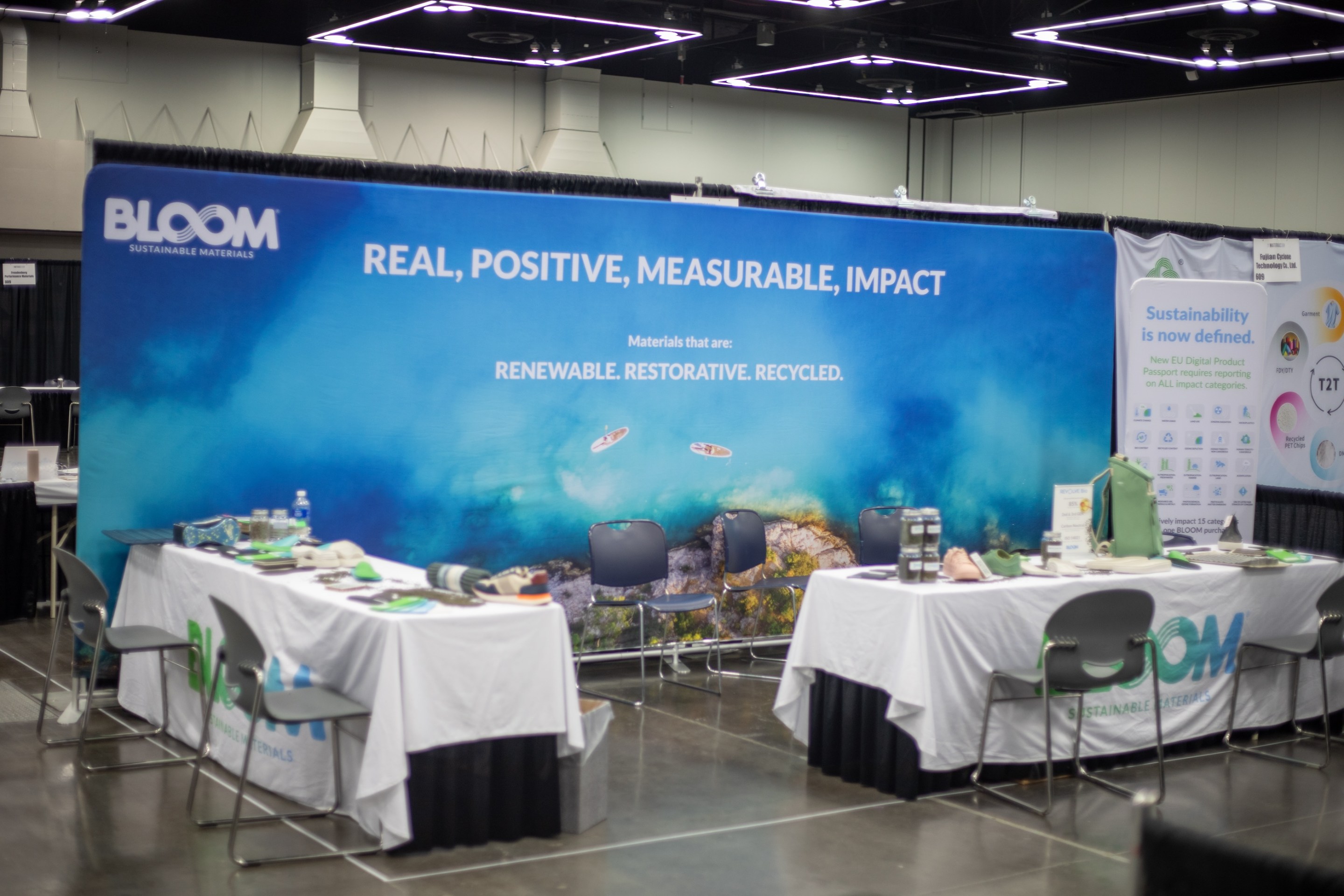 A booth at The Materials Show in Portland, Ore. in February of 2025. It's an empty couple of tables with a big banner behind it with the word BLOOM on it and some stuff about sustainability.