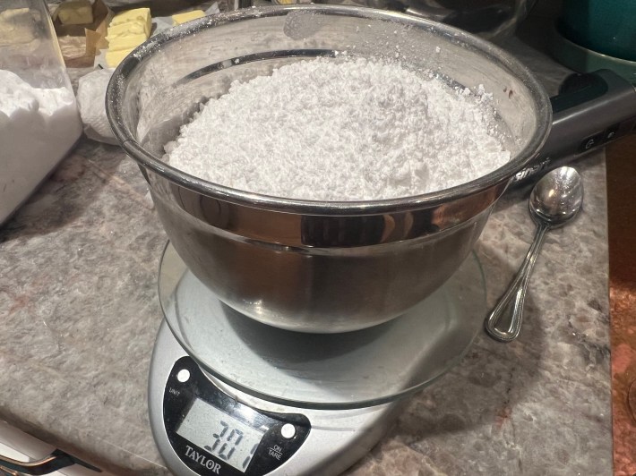 A bowl of powdered sugar sits on a food scale, showing a weight of 301 grams.