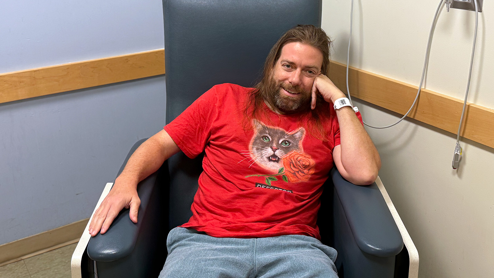 Dan McQuade sits in a blue chair, which is big and fake leather-ish, and has big arms on it. He has his head resting on his left arm, which is on the left arm of the chair. He has long light brown hair, a beard cropped to his face, and I dunno I'd say he's pretty handsome, but I am biased as I am the subject of the photo. Dan is wearing a red shirt with a grey and white tabby cat's face on it, with a rose under (and patially covering) the cat. Under that it says DEFECTOR with the 4 degrees slant logo. He has on blue shorts or pants (they're shorts, I know they're shorts), they're not jeans but they're not sweats, like a sort of inbetween situation. There's a hospital admit bracelet on his left arm. His right arm is on the other arm of the couch.