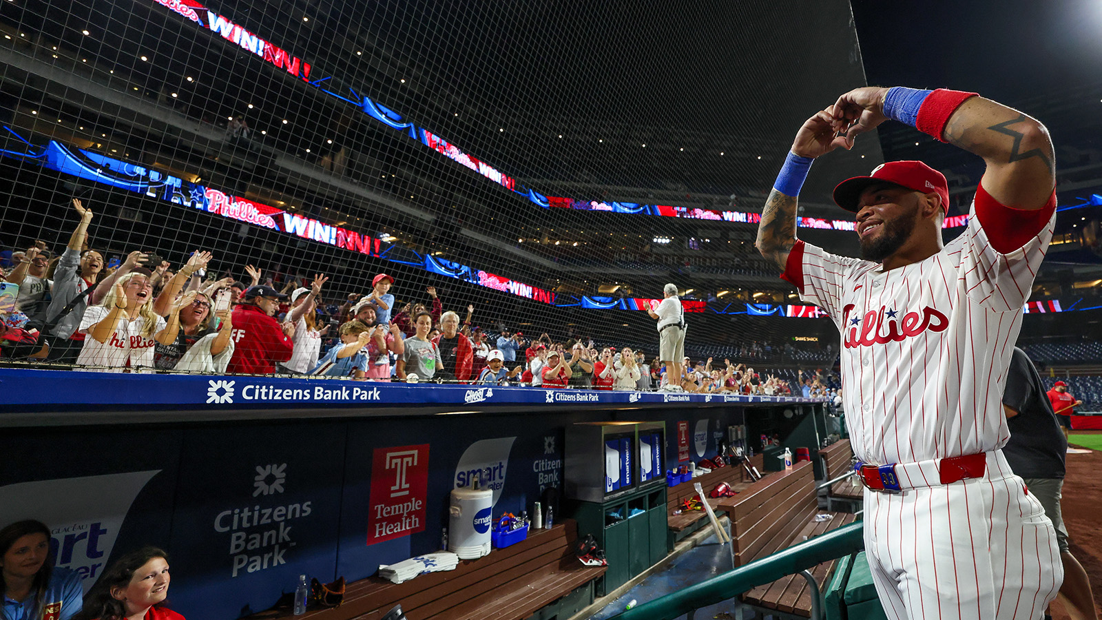Edmundo Sosa stands at the top of the dugout steps, making a heart with his hands above his head, as Phillies fans cheer. He's in his white pinstripe Phillies jersey.