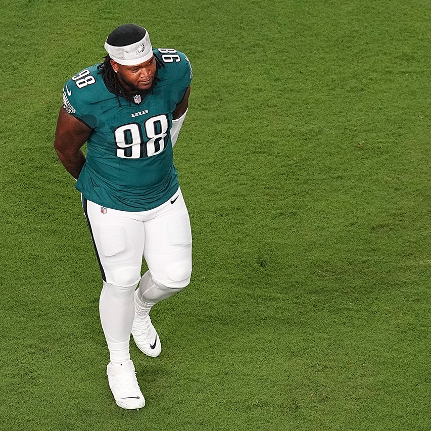 PHILADELPHIA, PENNSYLVANIA - SEPTEMBER 04: Jalen Carter #98 of the Philadelphia Eagles looks on after being ejected from the game against the Dallas Cowboys at Lincoln Financial Field on September 04, 2025 in Philadelphia, Pennsylvania. He's walking with his hands behind his back, in a midnight green uniform with white pants, his helmet off and a white bandana around his head. He's on the left side of the photo, and to the rest of him is green grass.