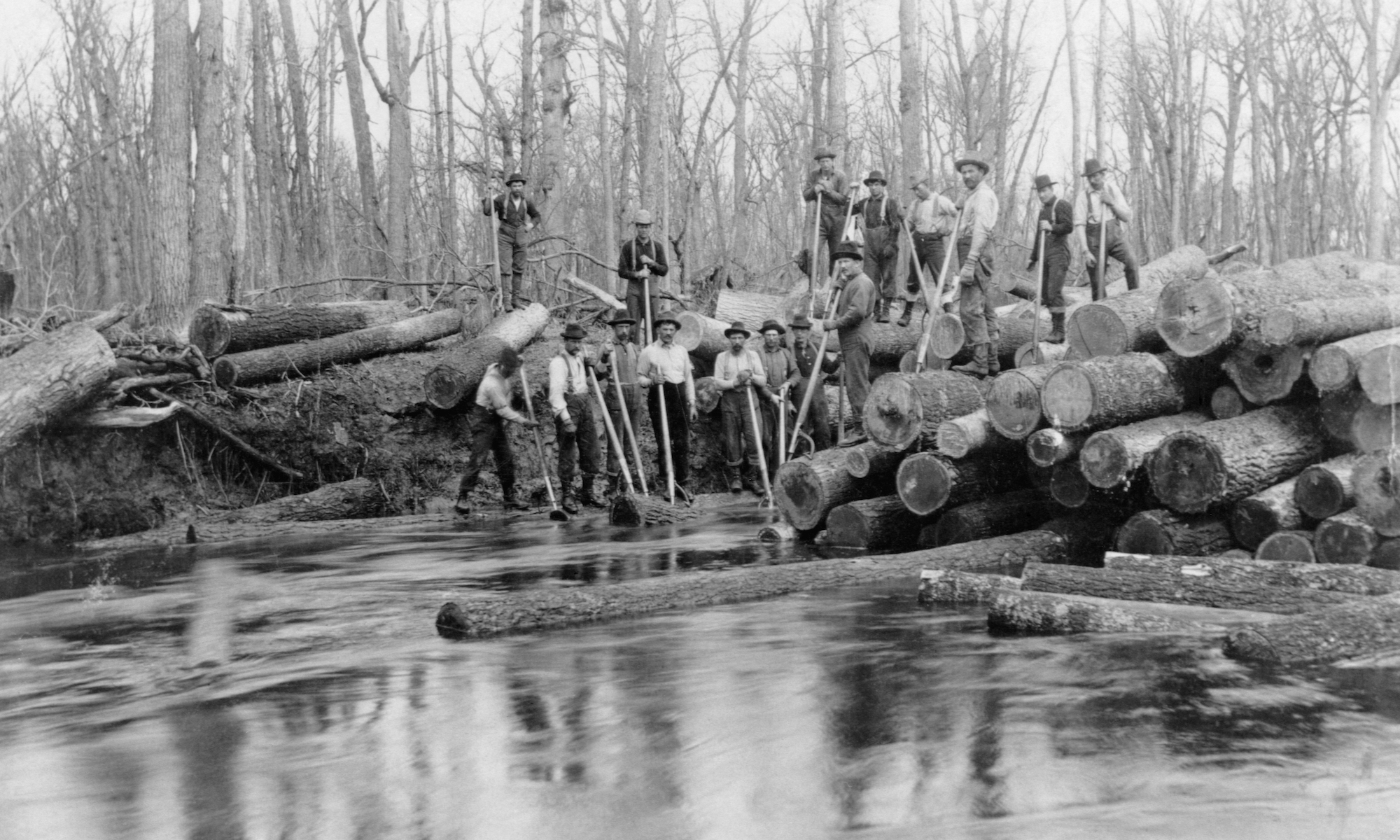 Old black-and-white photograph of log drivers along a Minnesota spillway. Fifteen or so grizzled men in work clothes and rumpled hats look up at the camera from across a river; they're using long poles to direct gigantic logs downstream.
