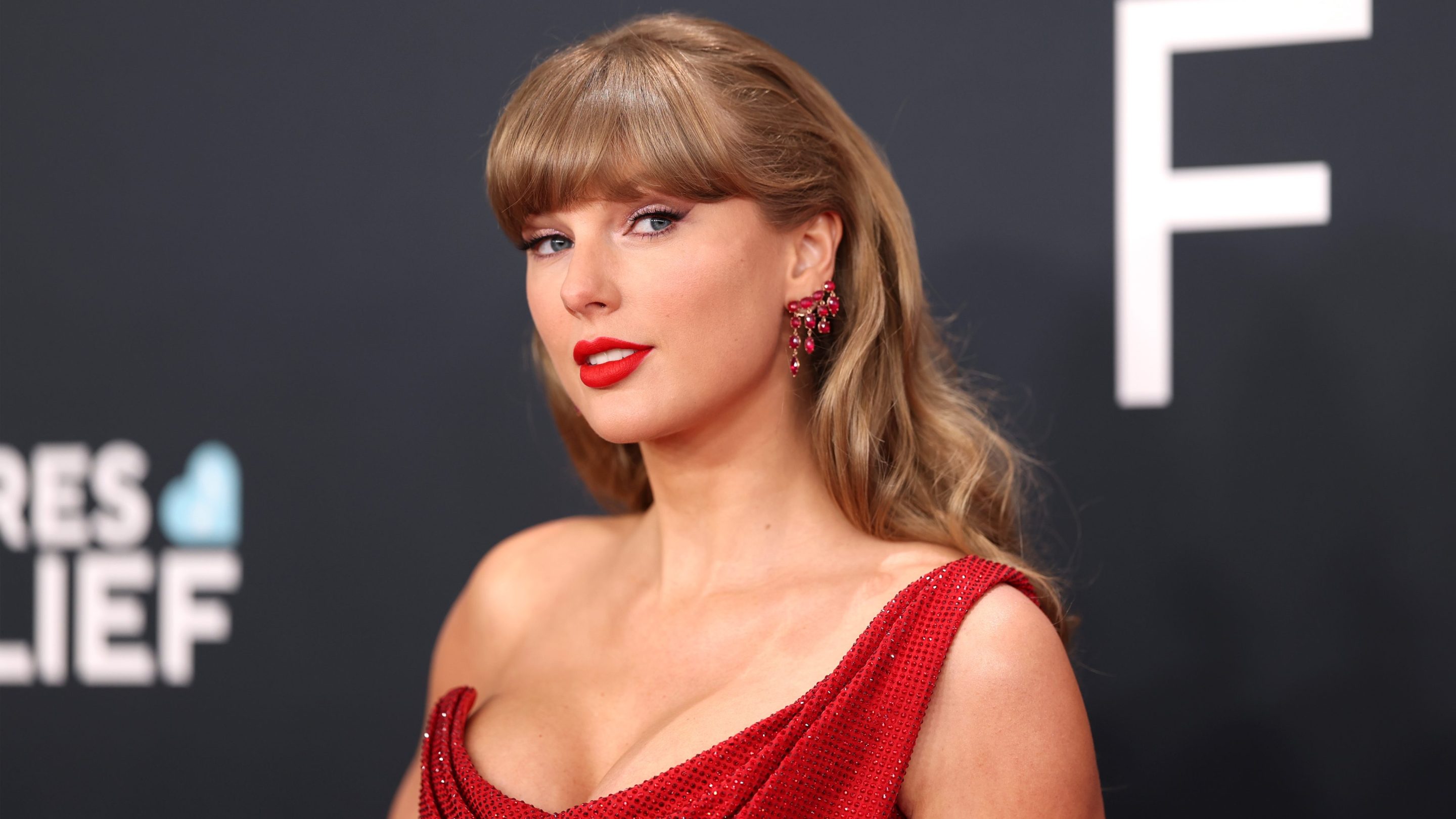 Taylor Swift on the red carpet wearing a red dress with winged eyeliner and looking out of the corner of her eye at the camera