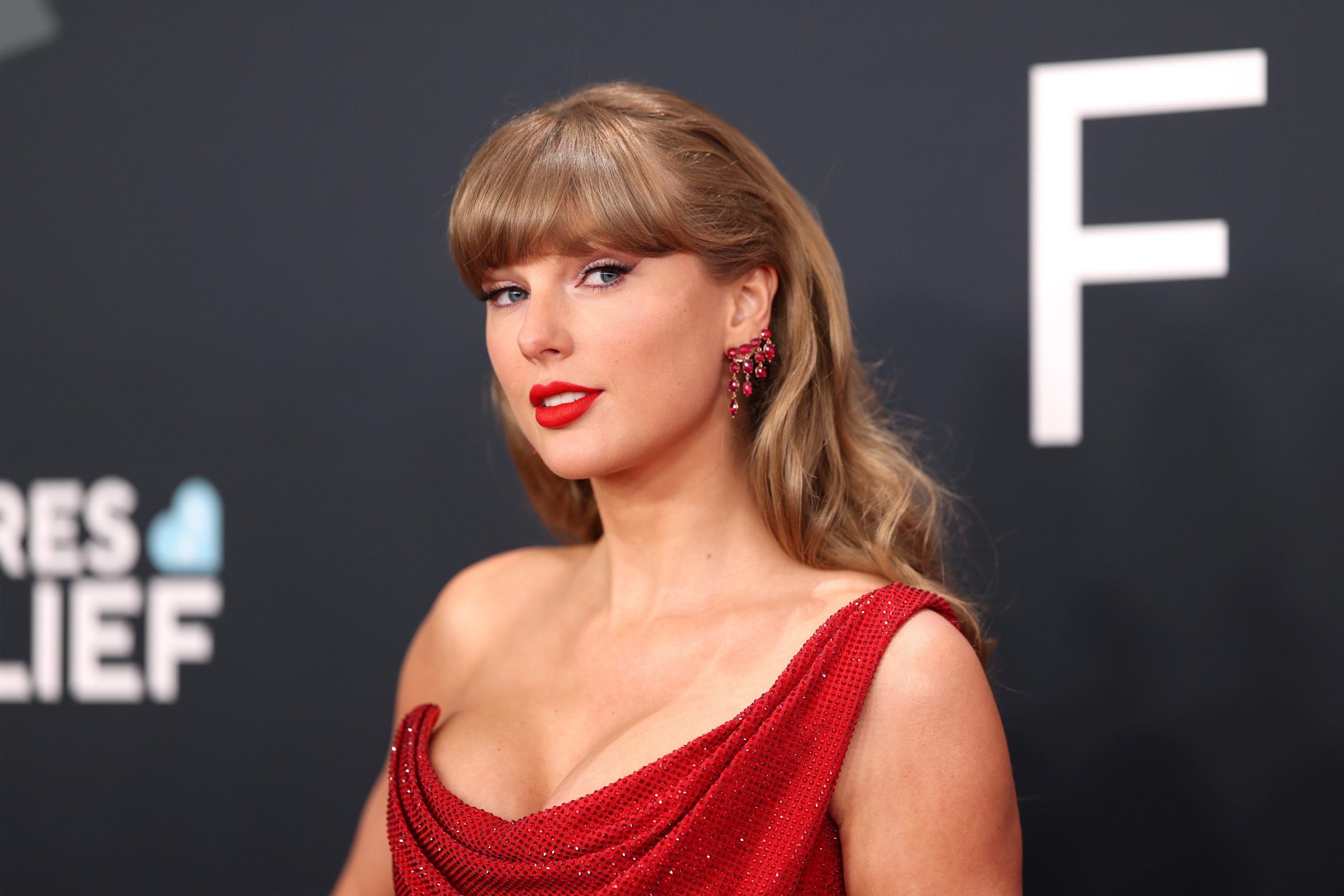 Taylor Swift on the red carpet wearing a red dress with winged eyeliner and looking out of the corner of her eye at the camera