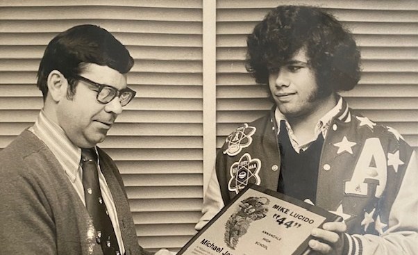 A photo of Mike Lucido having his jersey retired. He's wearing a letterman's jacket and sporting a huuuuge head of hear with massive sideburns. A man in a sweater and tie is handing him a plaque with his name on it and his now retired number, 44.