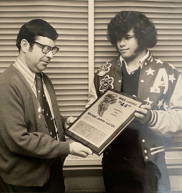 A photo of Mike Lucido having his jersey retired. He's wearing a letterman's jacket and sporting a huuuuge head of hear with massive sideburns. A man in a sweater and tie is handing him a plaque with his name on it and his now retired number, 44.